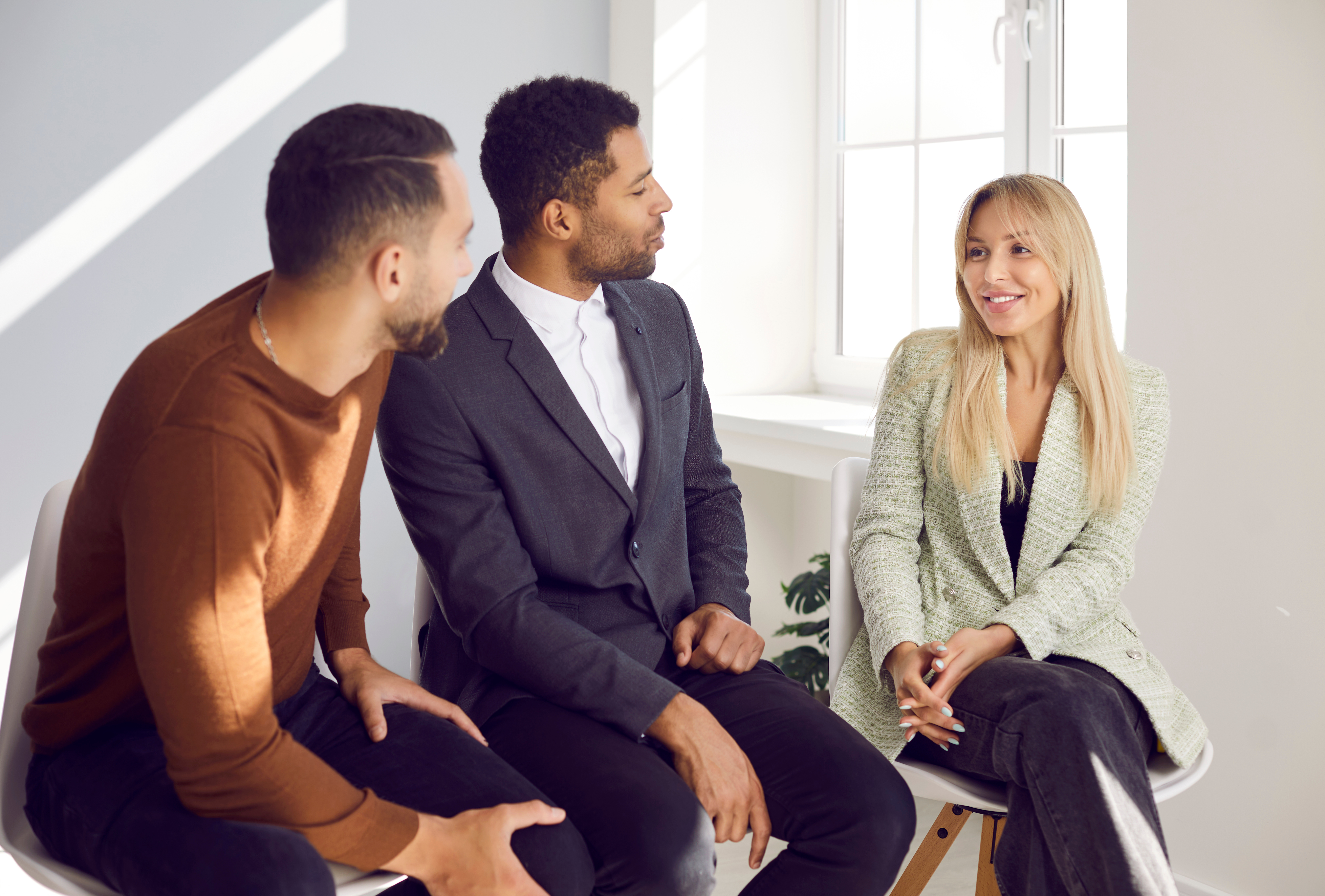 Colleagues have friendly conversation during informal meeting, exchange experiences and ideas. Two millennial men and women talk and discuss project while sitting on chairs in bright office.